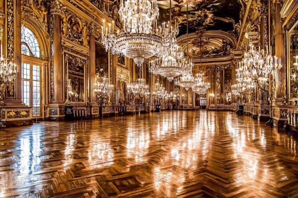 "Opulent ballroom photo with golden chandeliers, intricate carvings, and polished wooden floor."